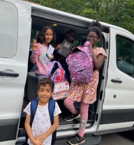 Three children children smile for the camera holding backpacks and school supplies.
